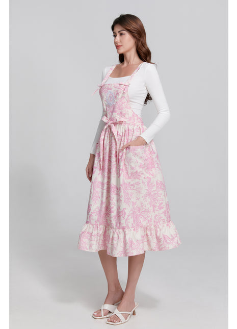 Model wearing a pink toile de jouy floral apron with pretty ruffle hemline on a gray background